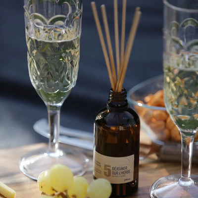 Diffuseur à bâtonnet naturel made in France en verre avec tiges à arômes, accompagné de raisins et de coupes de champagne